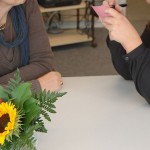 Eine ältere Dame und eine jüngere sitzen sich gegenüber, die junge notiert etwas auf einem Zettel. Im Vordergrund stehen Blumen.